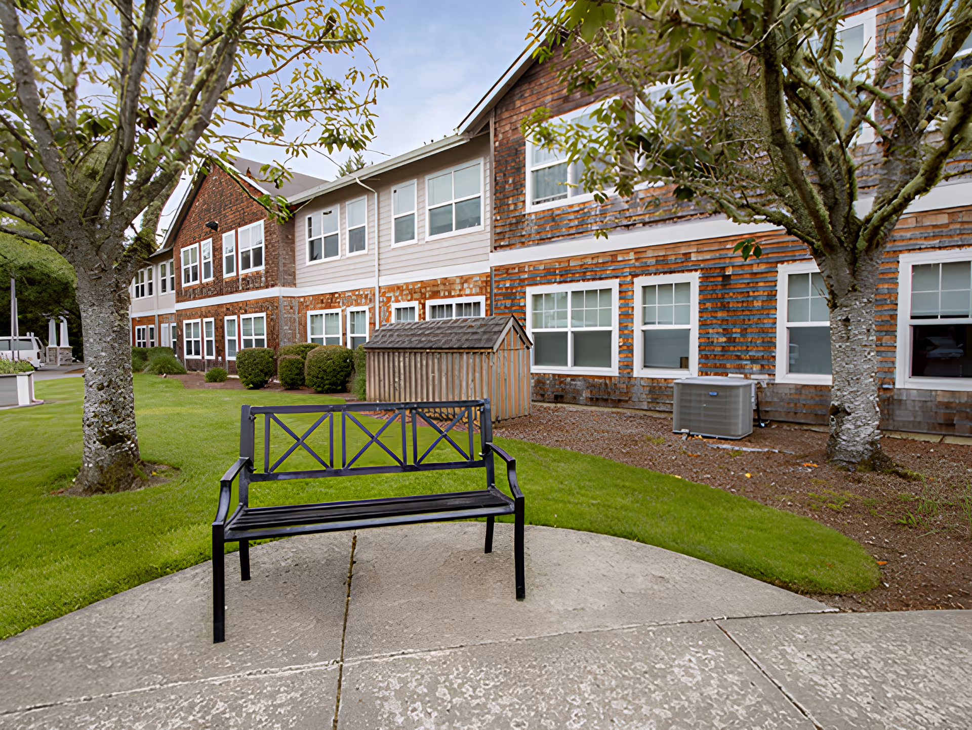 Outdoor area of Avamere at Seaside featuring a black metal bench on a concrete patio, green grass, two trees, and a two-story building with multiple windows and wooden siding in the background.
