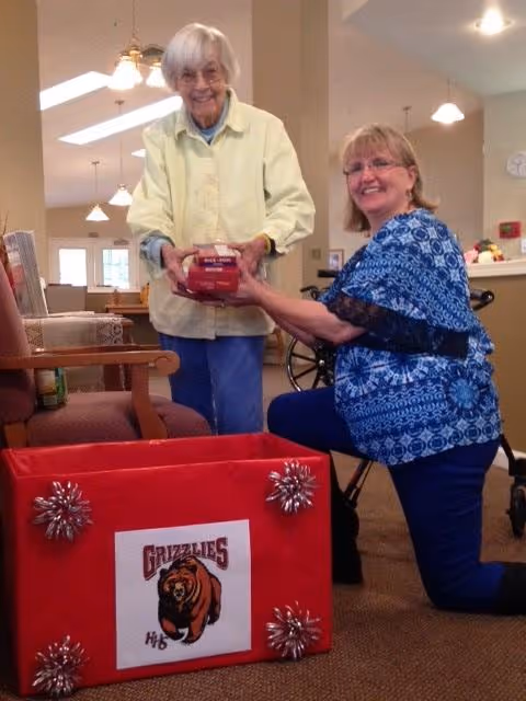 Two smiling elderly women in a communal living area place a wrapped package into a decorated red box labeled 'Grizzlies'.