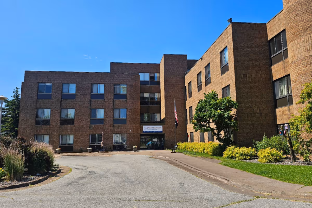 Front entrance of a multi-story brown brick senior living facility with an American flag and landscaped shrubs under a clear blue sky.