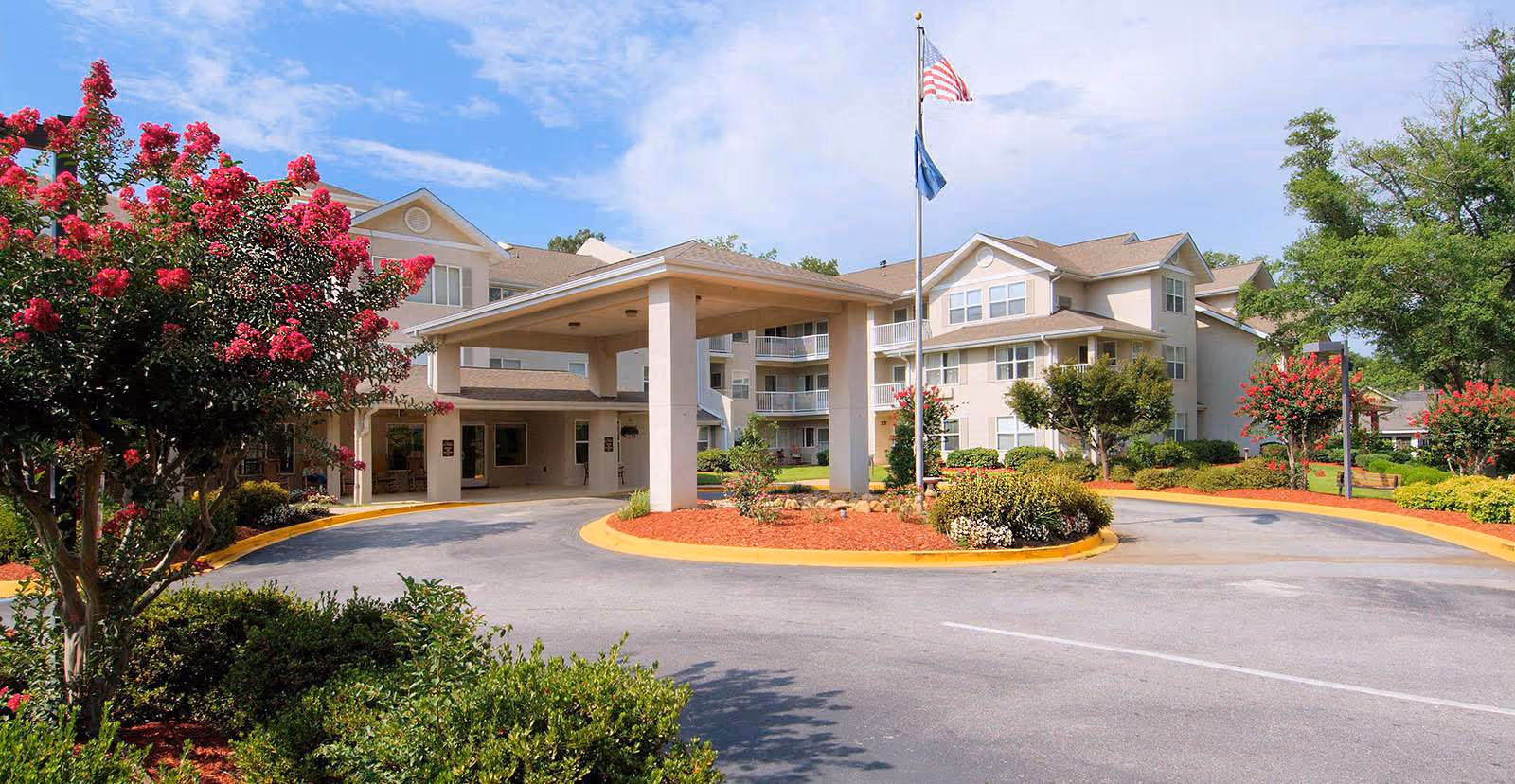 Exterior view of TerraBella Greenville senior living facility showing a three-story building with balconies, a covered entrance, landscaped gardens with flowering trees, and an American flag on a flagpole under a partly cloudy sky.