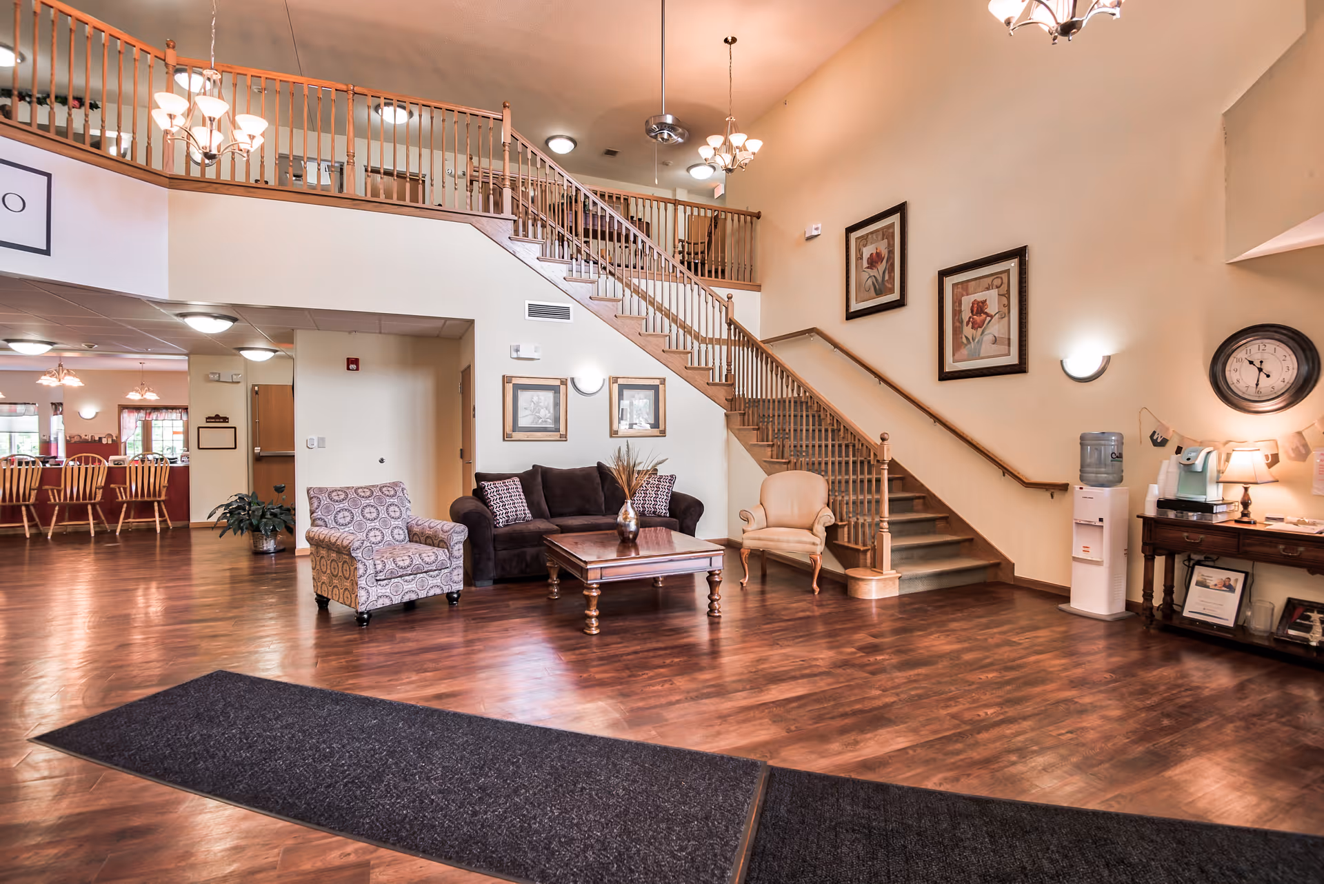 Spacious senior living lobby with seating area, wooden staircase and high ceilings leading to an upstairs balcony.