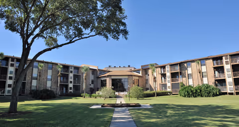 Exterior view of Brookdale Lake Tavares senior living facility showing a large, three-story building with balconies and windows surrounding a well-maintained grassy courtyard with a walkway, trees, and shrubs under a clear blue sky.