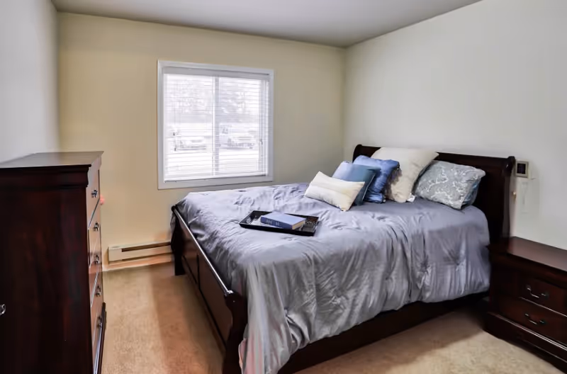 Bedroom with a made bed with gray bedding and pillows, a wooden dresser and nightstand, and a window with blinds.