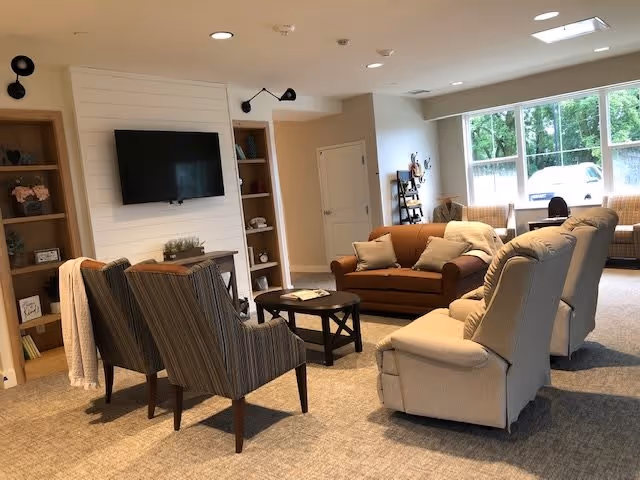 A cozy living room area with a mounted flat-screen TV on a white shiplap wall, flanked by built-in wooden shelves with decorative items. The room features a brown leather sofa, two beige recliners, and two striped armchairs around a round wooden coffee table. Large windows let in natural light, and the carpeted floor adds warmth to the space.