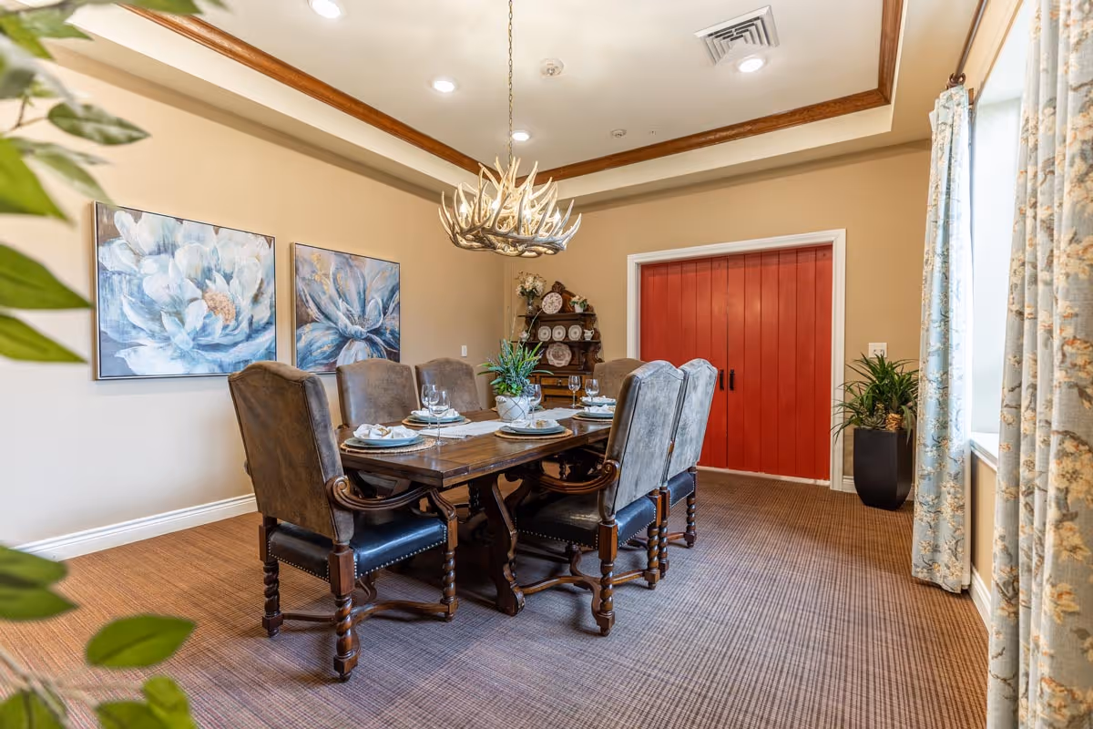 A dining room with a wooden table set for six people with plates, napkins, and glasses. The chairs are upholstered in a brownish-gray fabric with wooden legs. Above the table hangs a chandelier made of antlers. On the wall are two large floral paintings in shades of blue and white. There is a red double door at the far end of the room, a wooden cabinet with decorative plates, and a window with floral curtains letting in natural light. A potted plant is visible near the window.