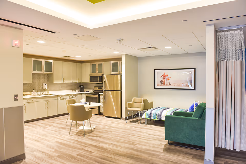 A bright and modern room featuring a kitchenette with beige cabinets, stainless steel refrigerator and oven, a small round dining table with two beige chairs, a green couch, a single bed with a colorful blanket, and a framed picture on the wall. There is also a privacy curtain on the right side.