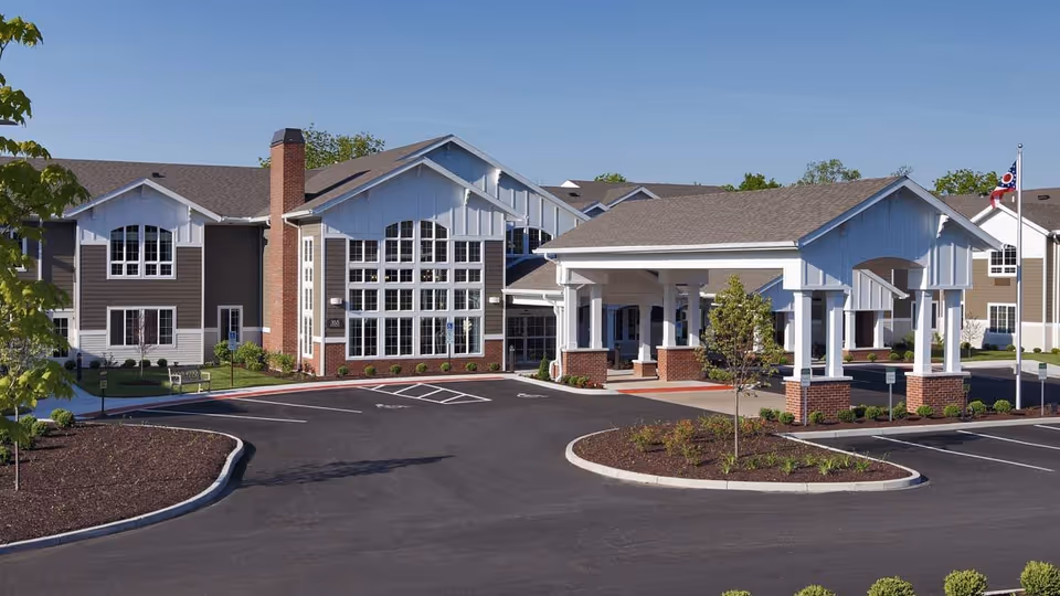Exterior view of The Enclave of Springboro senior living facility showing a large building with multiple windows, a covered entrance supported by white columns with brick bases, a parking lot, landscaped areas with small trees and shrubs, and a flagpole with the Ohio state flag.