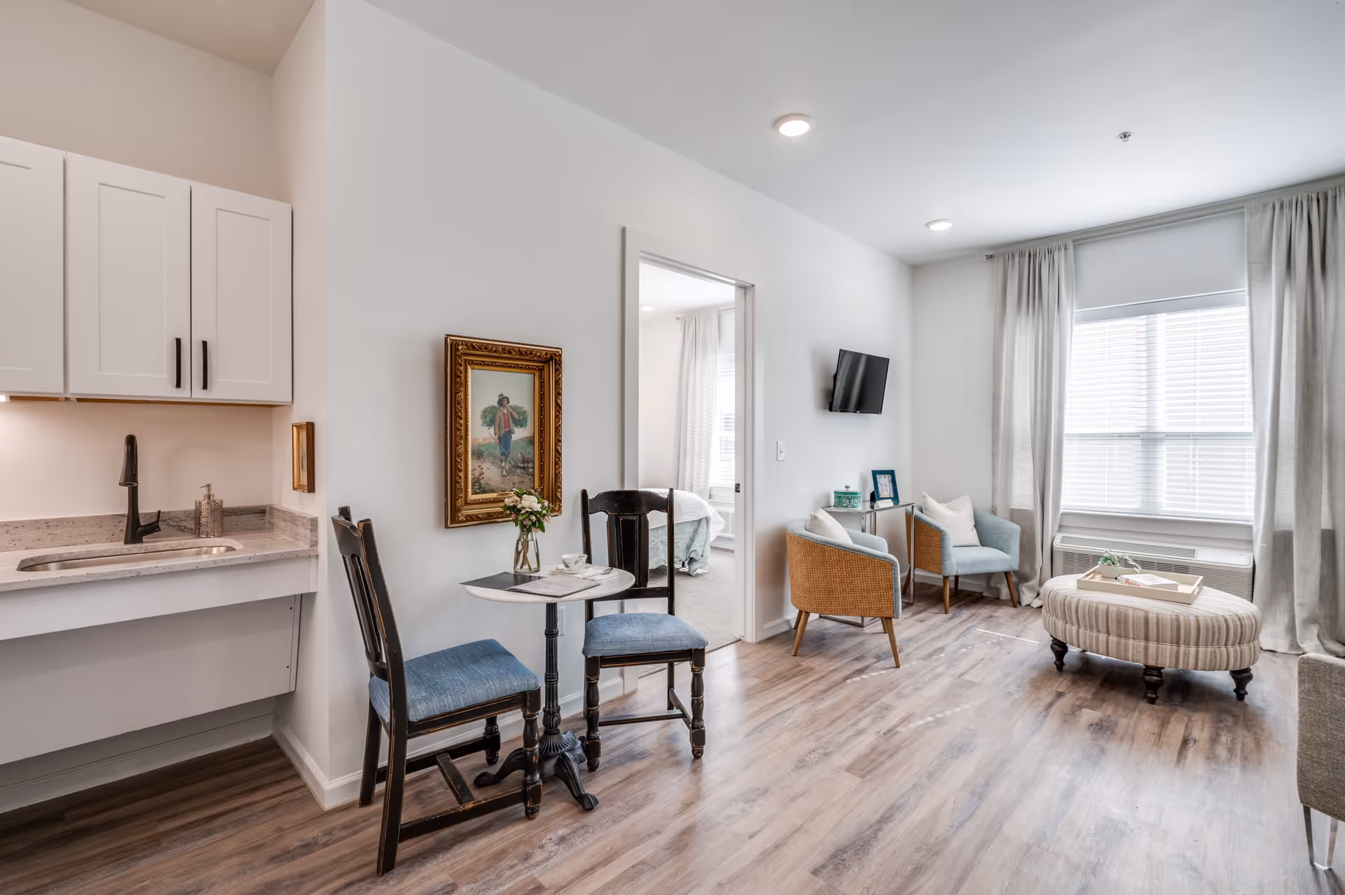 A bright and cozy living area in a senior living facility featuring a small kitchenette with white cabinets and a sink, a small round table with two chairs, a framed painting on the wall, two armchairs near a window with curtains, a wall-mounted TV, and a striped ottoman on wooden flooring. A bedroom with a bed is visible through an open doorway.