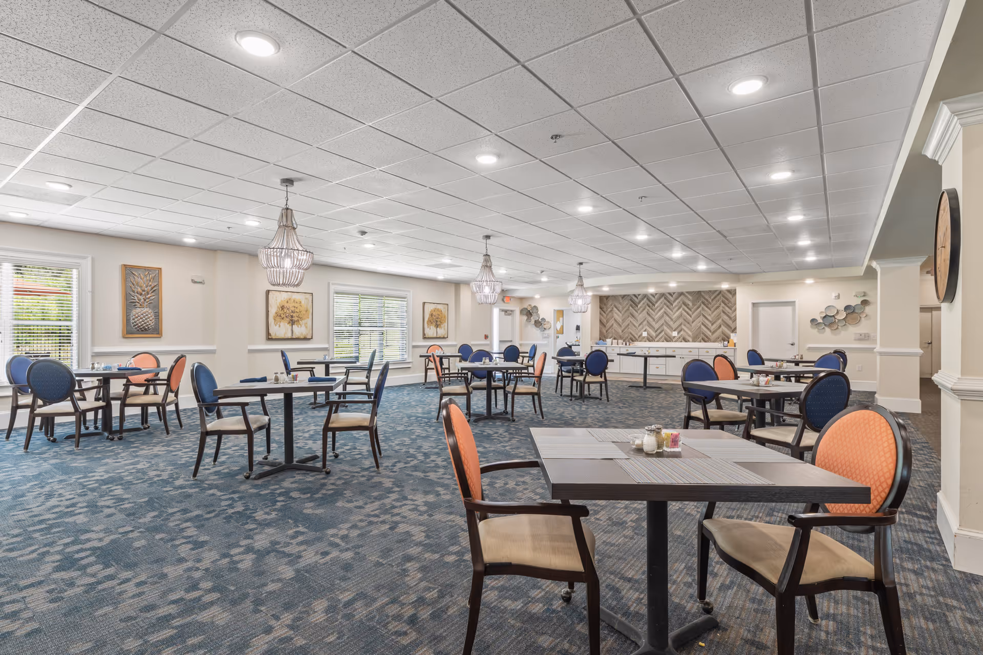 A spacious dining room with multiple tables and chairs arranged neatly. The chairs have blue and orange upholstery. The room features large windows with blinds, decorative wall art, and three hanging chandeliers. The carpet is patterned in shades of blue, and the walls are painted a light cream color. There is a counter with cabinets along the back wall.