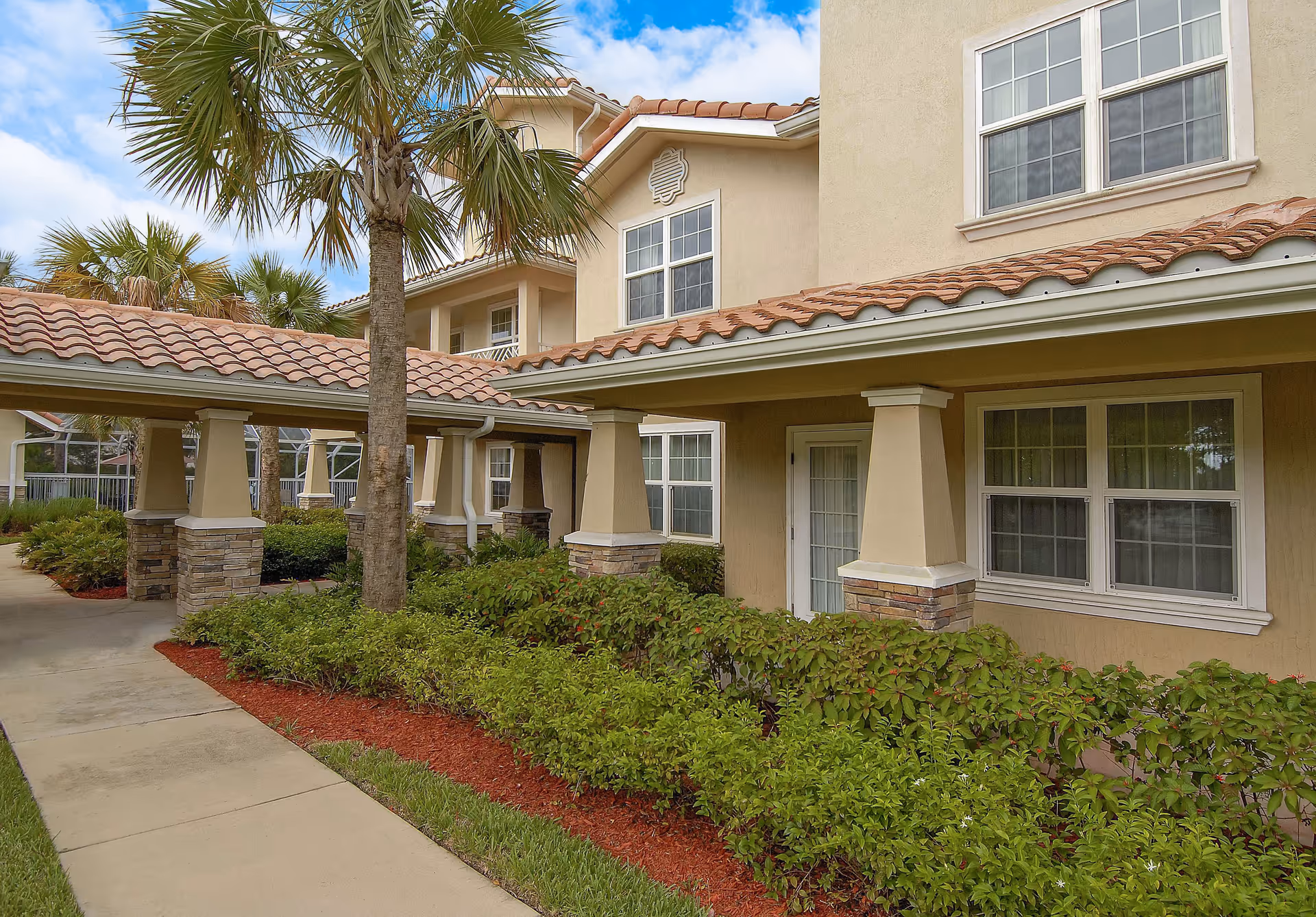 Exterior view of a beige stucco building with a tiled roof, covered walkways, palm trees, and landscaped shrubs.