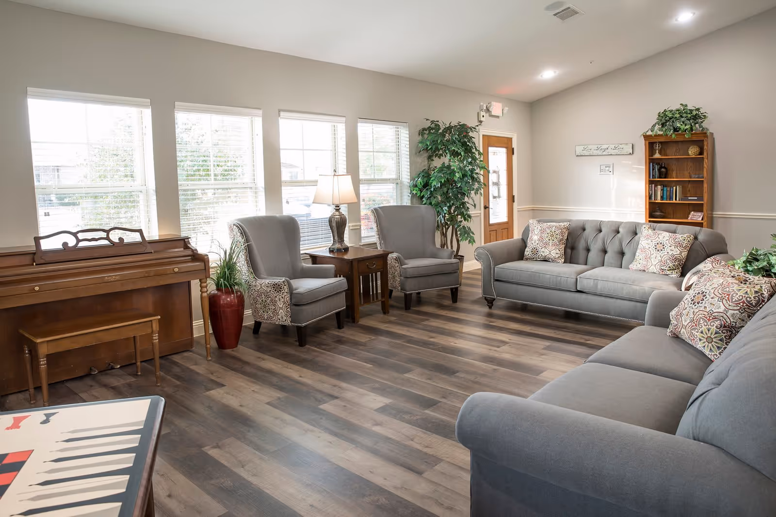 A bright and spacious living room with large windows letting in natural light. The room features two gray sofas with patterned throw pillows, two gray armchairs with patterned sides, a wooden piano with a matching bench, a wooden side table with a lamp, a tall green plant in a red pot, and a wooden bookshelf filled with books and decorative items. The floor is covered with dark wood laminate, and a door with glass panels is visible in the background.