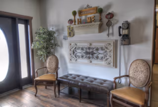 A cozy seating area near an entrance door with two upholstered wooden chairs and a tufted leather bench. The wall behind features decorative elements including a wooden shelf with the word 'HOME', ornamental wall art, and a vintage wall-mounted telephone. A potted plant is placed next to the door.