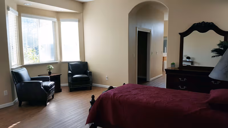 A bedroom in an assisted living facility with a bed covered in a burgundy blanket, two black leather armchairs near a bay window, a wooden dresser with a large mirror, and a small table with a plant. The room has light-colored walls and wood flooring.