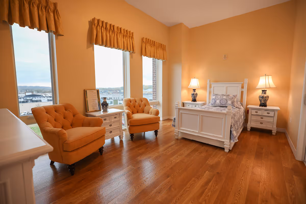 A bright bedroom with wooden flooring featuring a white wooden bed with blue and white bedding, two matching white nightstands with blue and white lamps, and two orange upholstered armchairs near large windows with orange valances overlooking a cityscape.