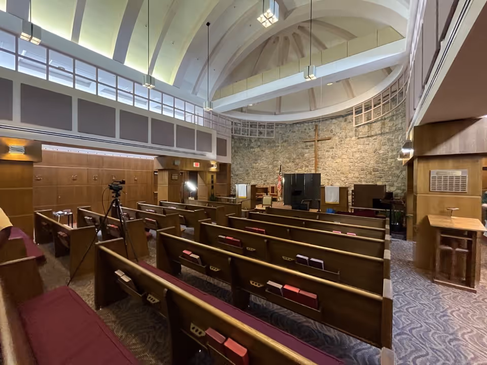 Interior view of a chapel or worship room with wooden pews, a stone wall with a wooden cross, an American flag, and a high vaulted ceiling with hanging lights. There is a tripod with a camera set up among the pews.