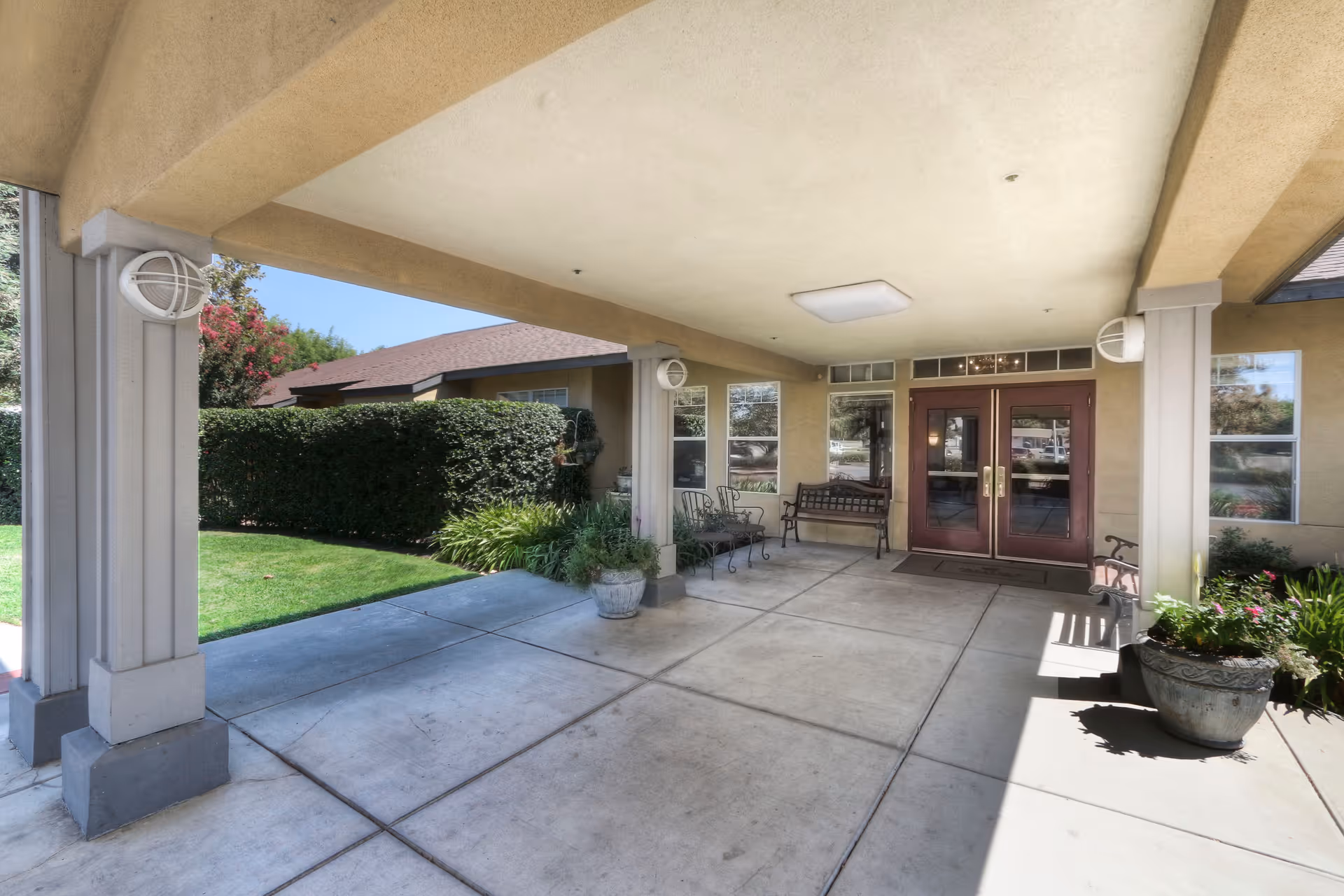 Covered entrance portico with seating, potted plants, and double glass doors leading into the building.