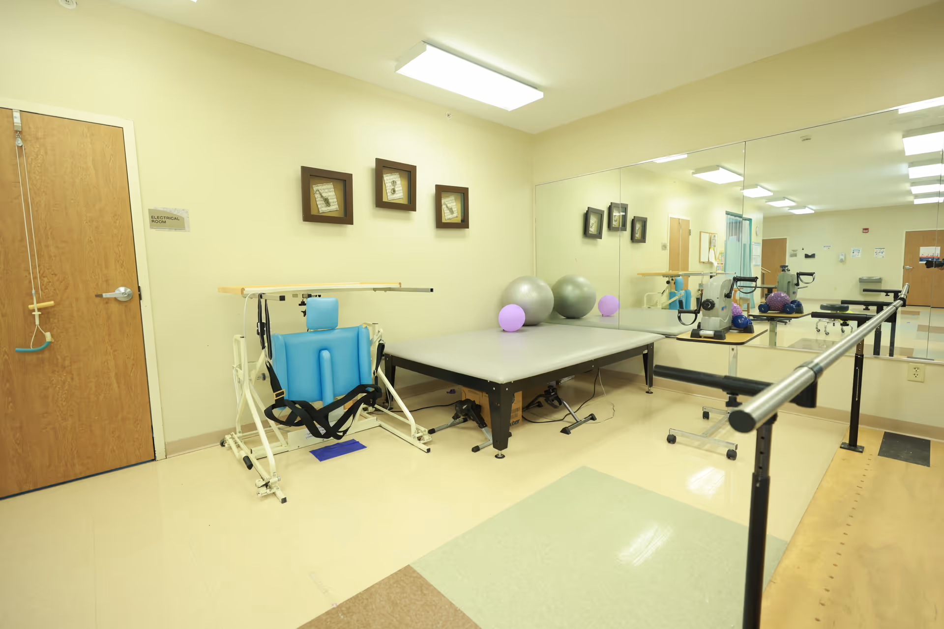 A physical therapy room with a blue rehabilitation chair, a padded exercise table with exercise balls and balloons on it, a parallel bar for walking exercises, and a large wall mirror reflecting the room.