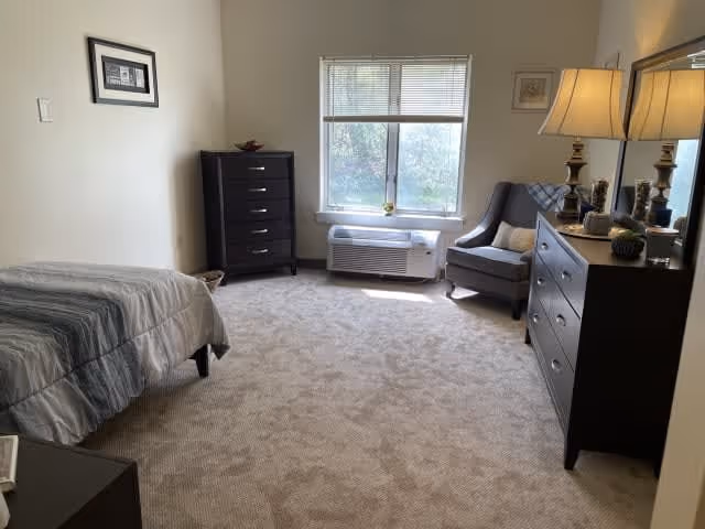 A bright and clean bedroom with a single bed covered in a gray striped comforter on the left, a tall dark dresser against the far wall, a window with blinds letting in natural light, a gray upholstered armchair with a pillow, and a dark dresser with a lamp and decorative items on the right.