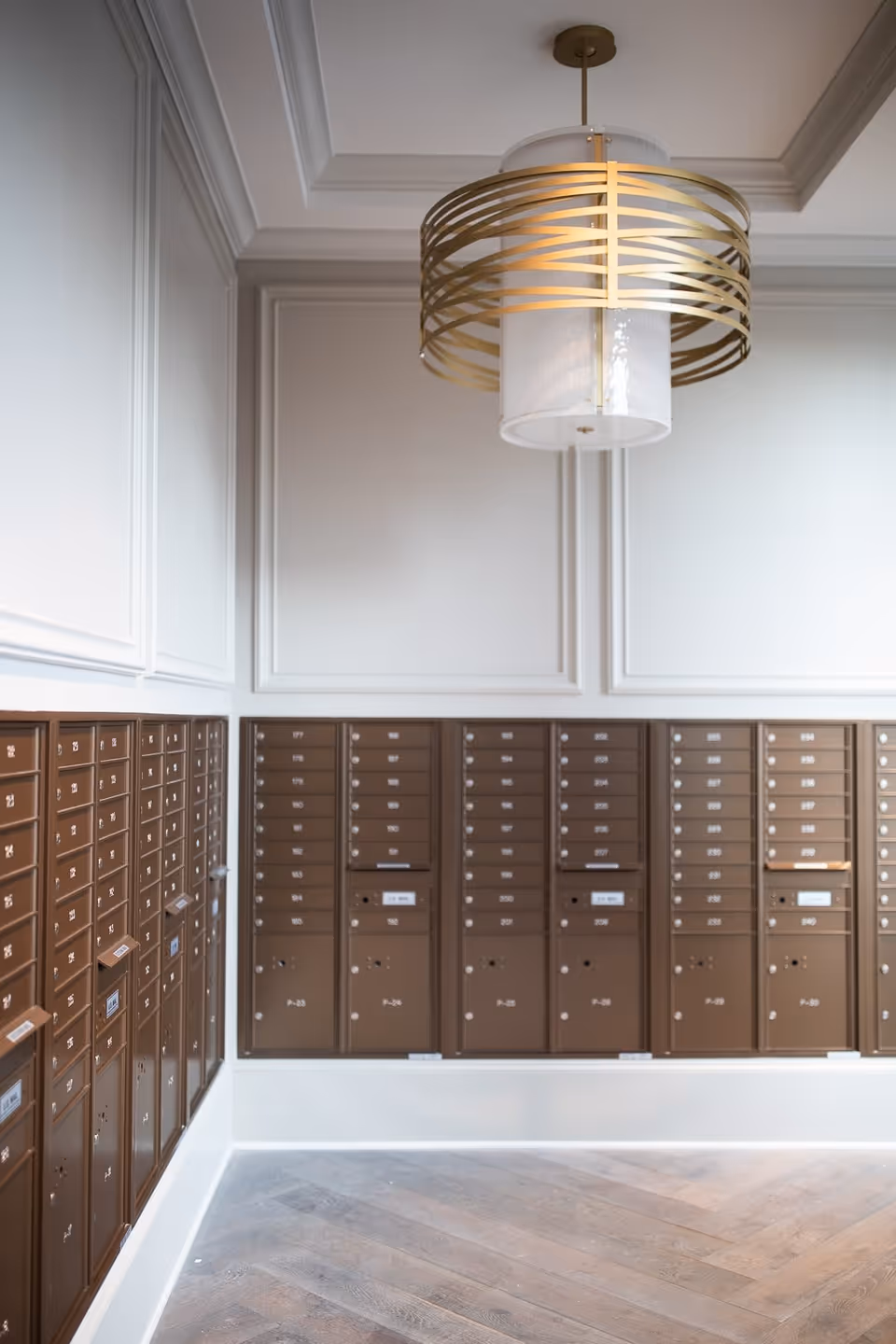 Interior view of a mailroom with multiple brown mailboxes mounted on white paneled walls and a modern gold and white ceiling light fixture above a wooden floor.