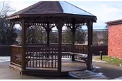 Wooden octagonal gazebo in an outdoor courtyard next to a brick building with trees and a walkway.