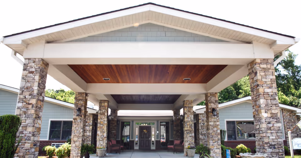 Entrance of The Village At Allandale facility showing a covered driveway supported by stone pillars with seating areas on either side and greenery around the building.