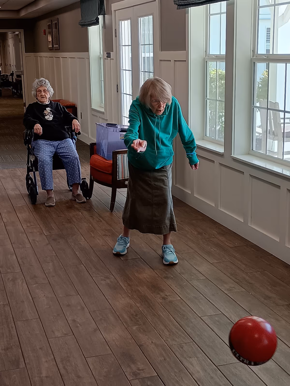 Two elderly women in a hallway with wooden flooring and large windows. One woman in a green hoodie and skirt is rolling a red ball on the floor, while the other woman sits in a wheelchair watching her. The hallway has white paneled walls and natural light coming through the windows.