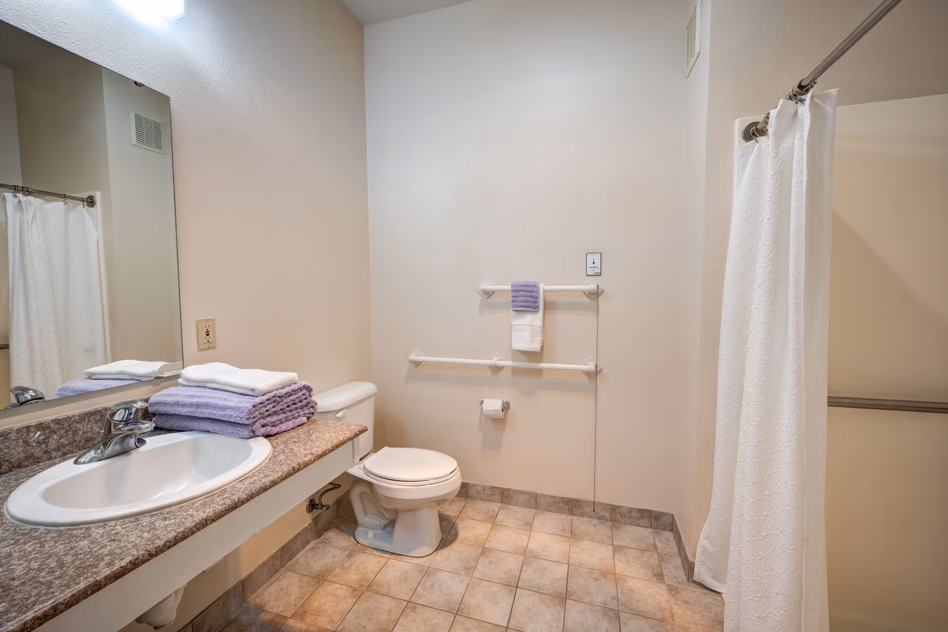 Accessible bathroom with a sink and countertop, toilet with grab bars, and a shower behind a white curtain.