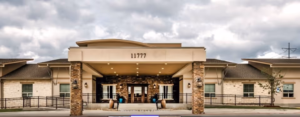 Front exterior view of La Fontaine Memory Care facility showing a single-story building with stone and beige walls, a covered entrance supported by stone pillars, and the address number 11777 displayed above the entrance.