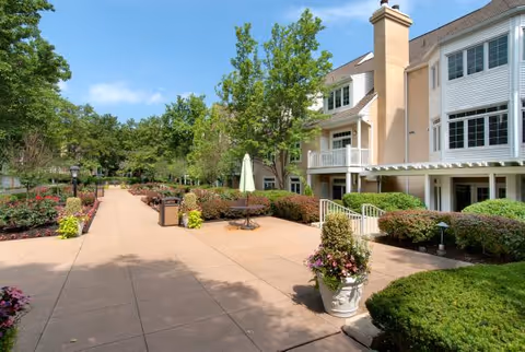 Outdoor patio area at Evergreen Retirement Community with paved walkways, landscaped bushes, trees, flower pots, and a building with balconies and large windows under a clear blue sky.