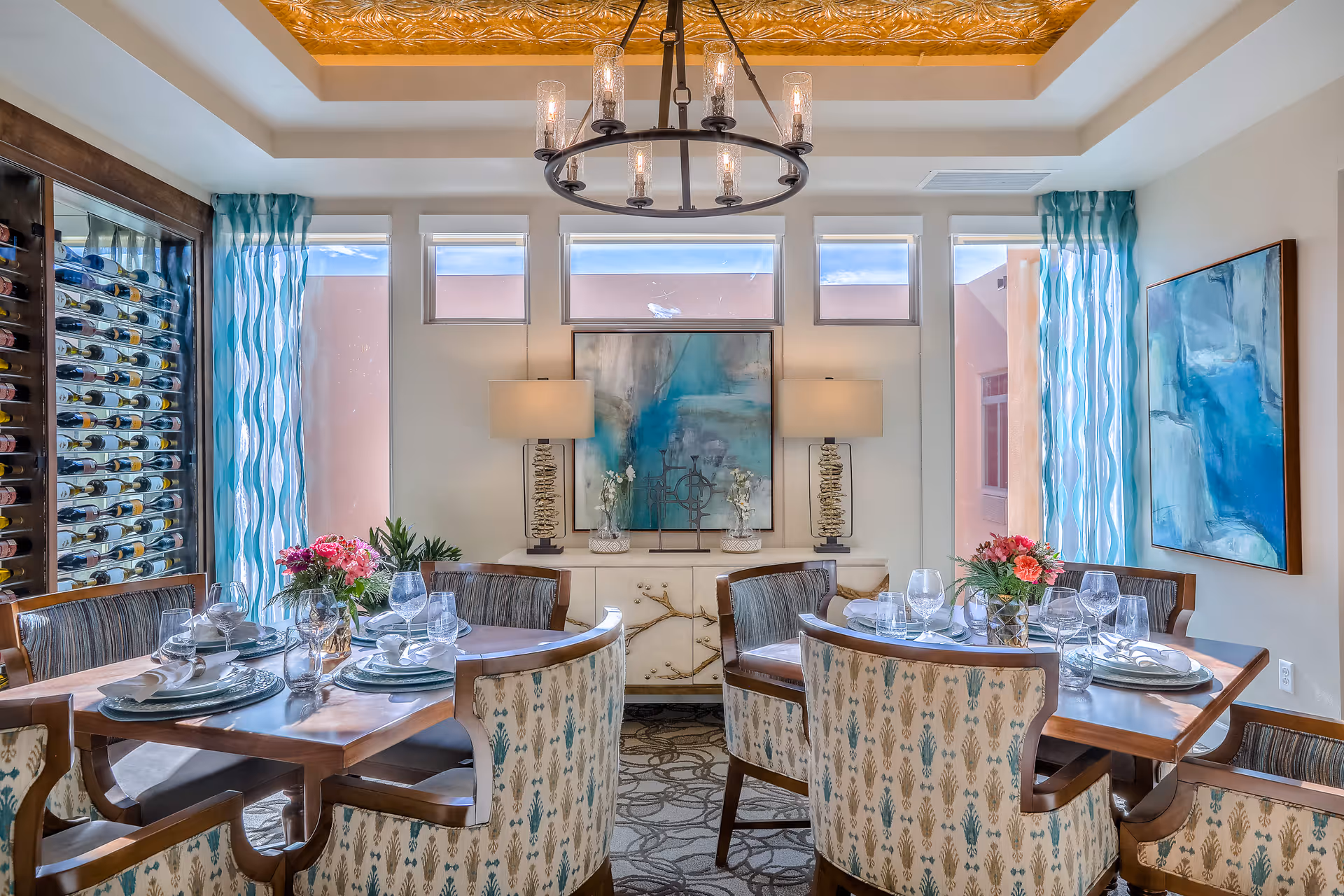A bright and elegant dining room with two wooden tables set for a meal, each surrounded by upholstered chairs with patterned fabric. The room features large windows with blue patterned curtains, a wine rack filled with bottles on the left, and a sideboard with two lamps and decorative items beneath a large abstract painting. A modern chandelier hangs from a ceiling with a golden decorative inset.