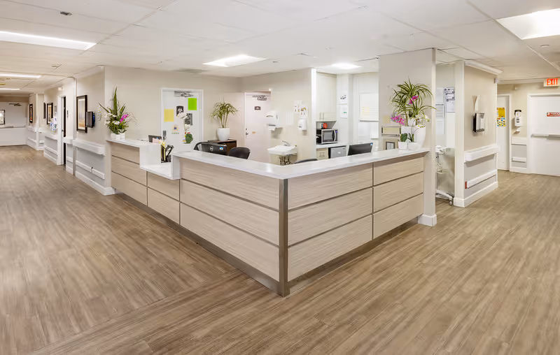 A clean and well-lit reception desk area in a senior living facility with light wood paneling and white countertops. The space features wood-look flooring, white walls, potted plants on the counter, and doors leading to other rooms. The ceiling has recessed lighting and there are handrails along the walls in the hallway.