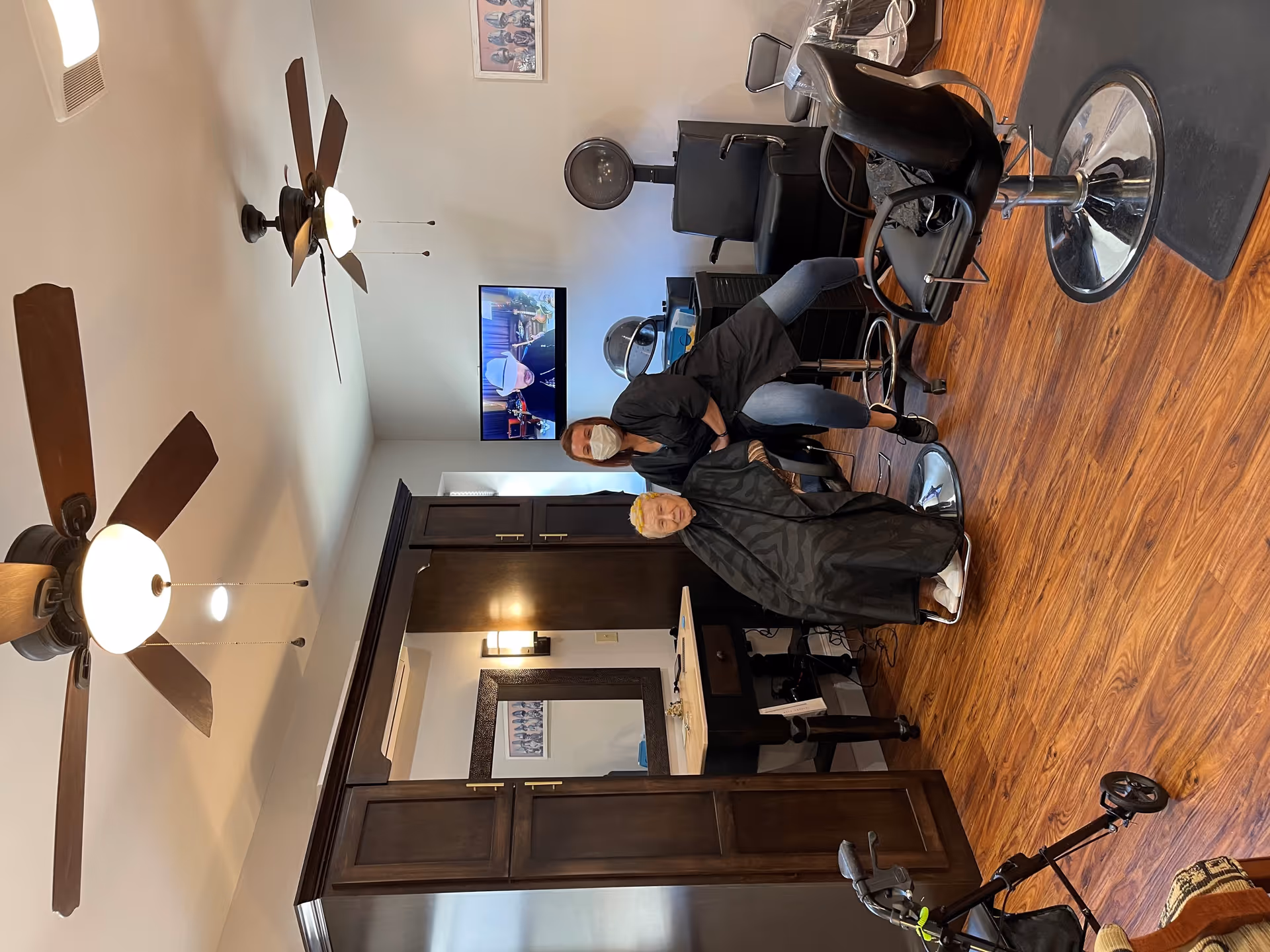 A hair salon area inside a senior living facility with wooden floors and ceiling fans. A hairstylist wearing a mask is standing behind an elderly person seated in a salon chair with a black cape. There are salon equipment and chairs around, a large mirror on the wall, and a TV mounted on the wall showing a person wearing a mask.