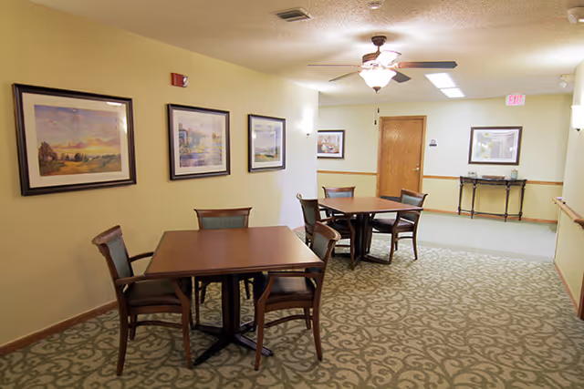 A small communal dining area with two square wooden tables and chairs, framed landscape paintings on light yellow walls, and patterned carpet.
