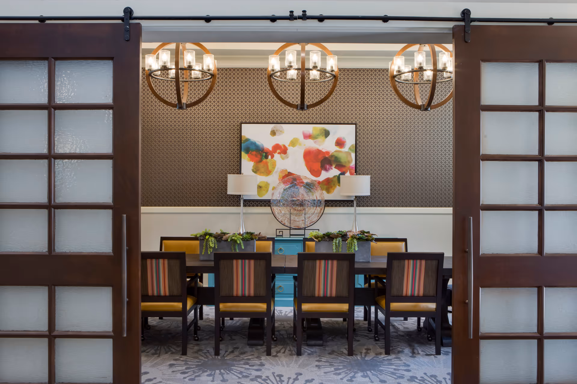 A dining room with a long dark wooden table surrounded by eight chairs with yellow seats and striped backs. The room features two modern table lamps on a blue sideboard against a patterned wallpapered wall. Above the sideboard is a colorful abstract painting. Three circular chandeliers hang from the ceiling. The room is viewed through partially open sliding wooden doors with frosted glass panels.