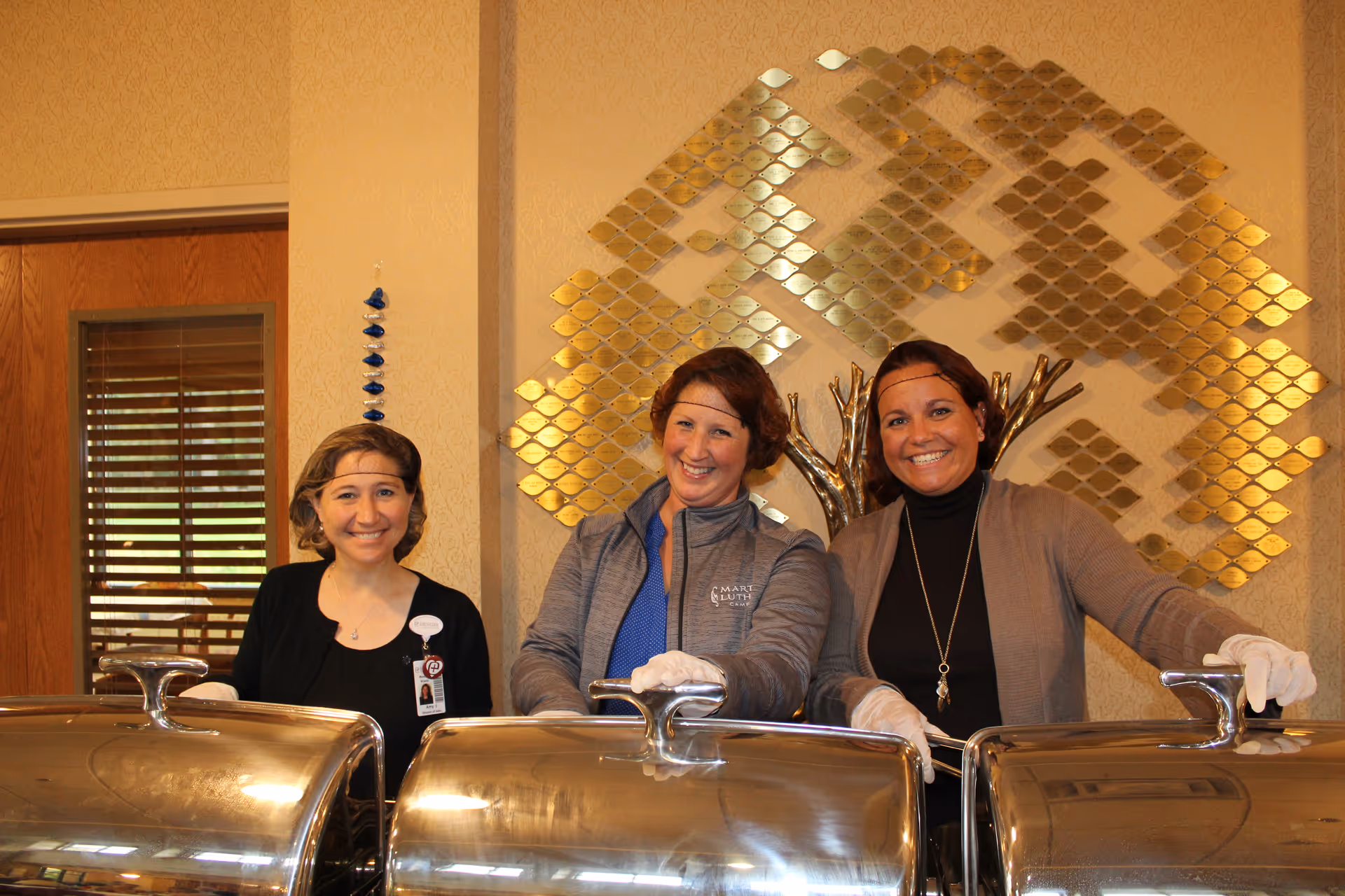 Three smiling staff members stand behind metal chafing dishes in a dining/service area.