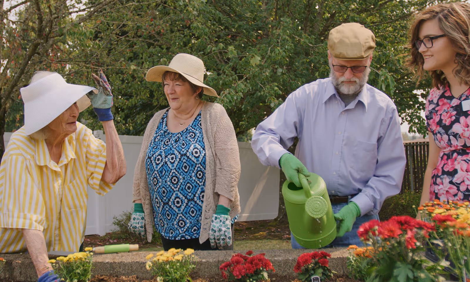 Four people gardening outdoors near a flower bed. Three seniors, two women and one man, are wearing gardening gloves and hats. The man is watering flowers with a green watering can. A younger woman in a floral dress is standing nearby, smiling. Trees and a white fence are in the background.