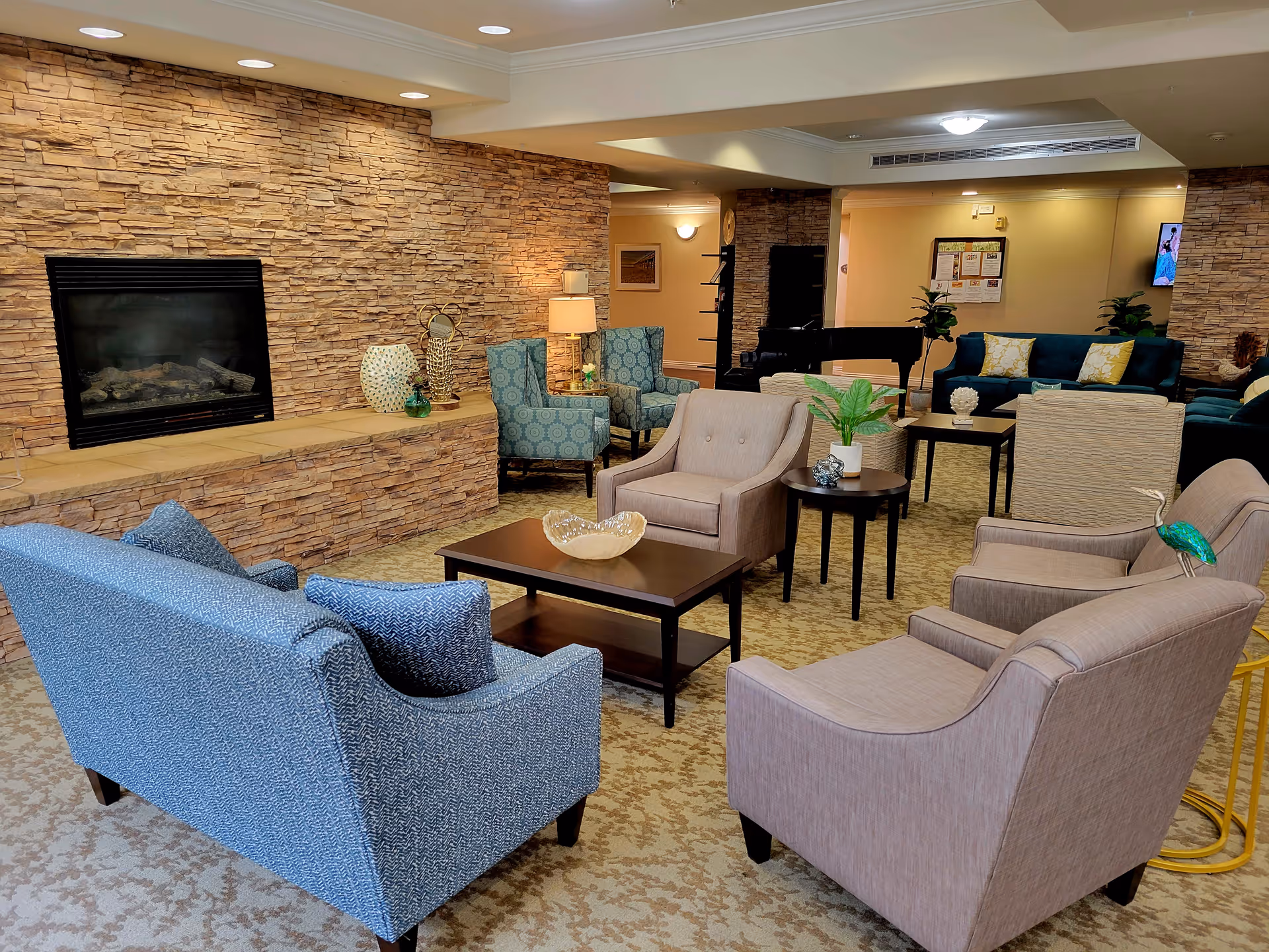 A cozy senior living lounge with upholstered chairs and sofas arranged around a coffee table in front of a stone fireplace.
