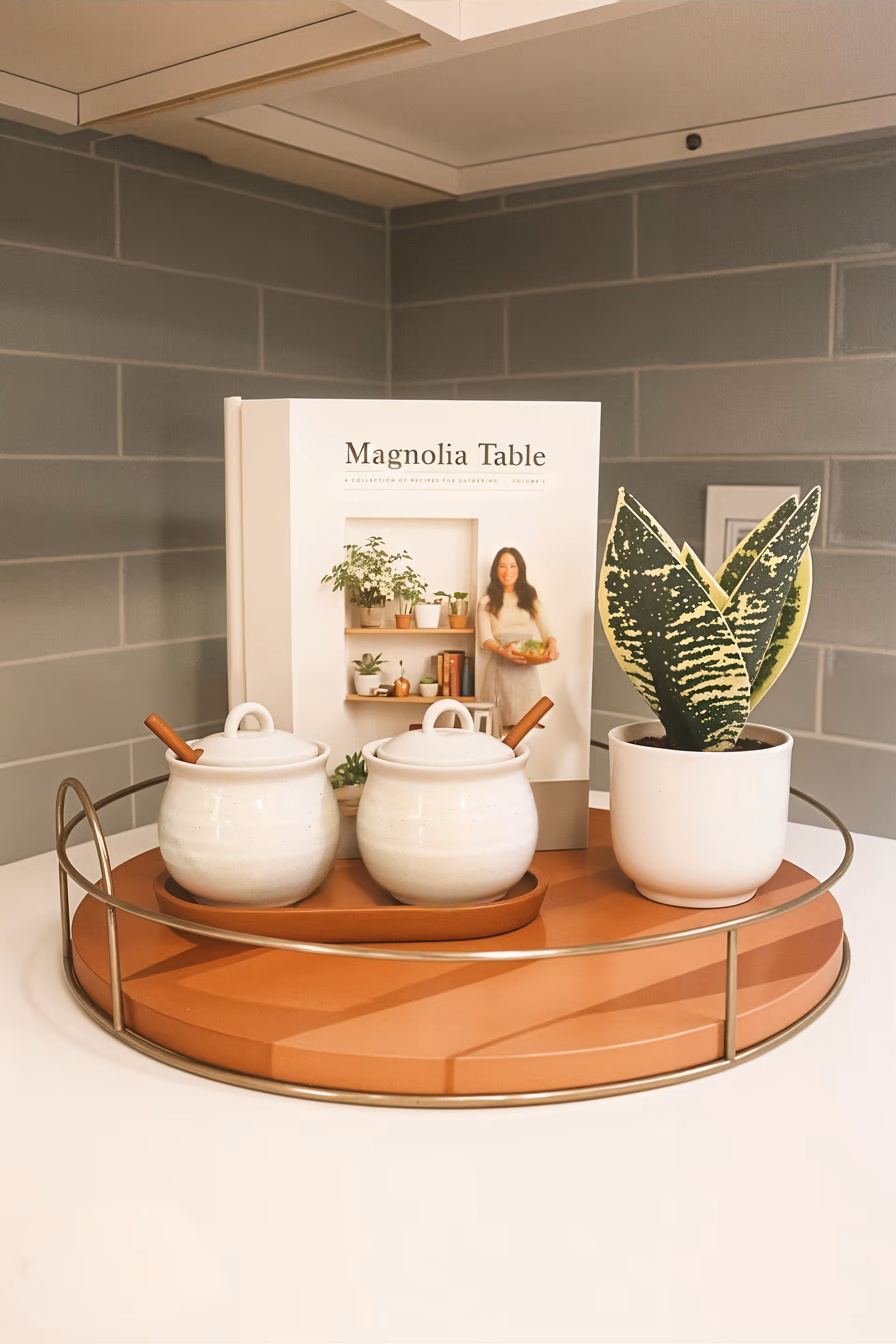 A decorative tray on a white surface holding two white ceramic jars with wooden spoons, a potted snake plant, and a book titled 'Magnolia Table' featuring a woman holding a bowl on the cover. The background shows a gray tiled wall.