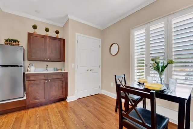 A small dining area with a dark wooden table set for two with yellow plates and glasses, next to a window with white plantation shutters. To the left, there is a kitchenette with dark wooden cabinets, a small sink, and a stainless steel mini refrigerator. The floor is hardwood, and the walls are painted beige with white trim. A round mirror hangs on the wall near the window.