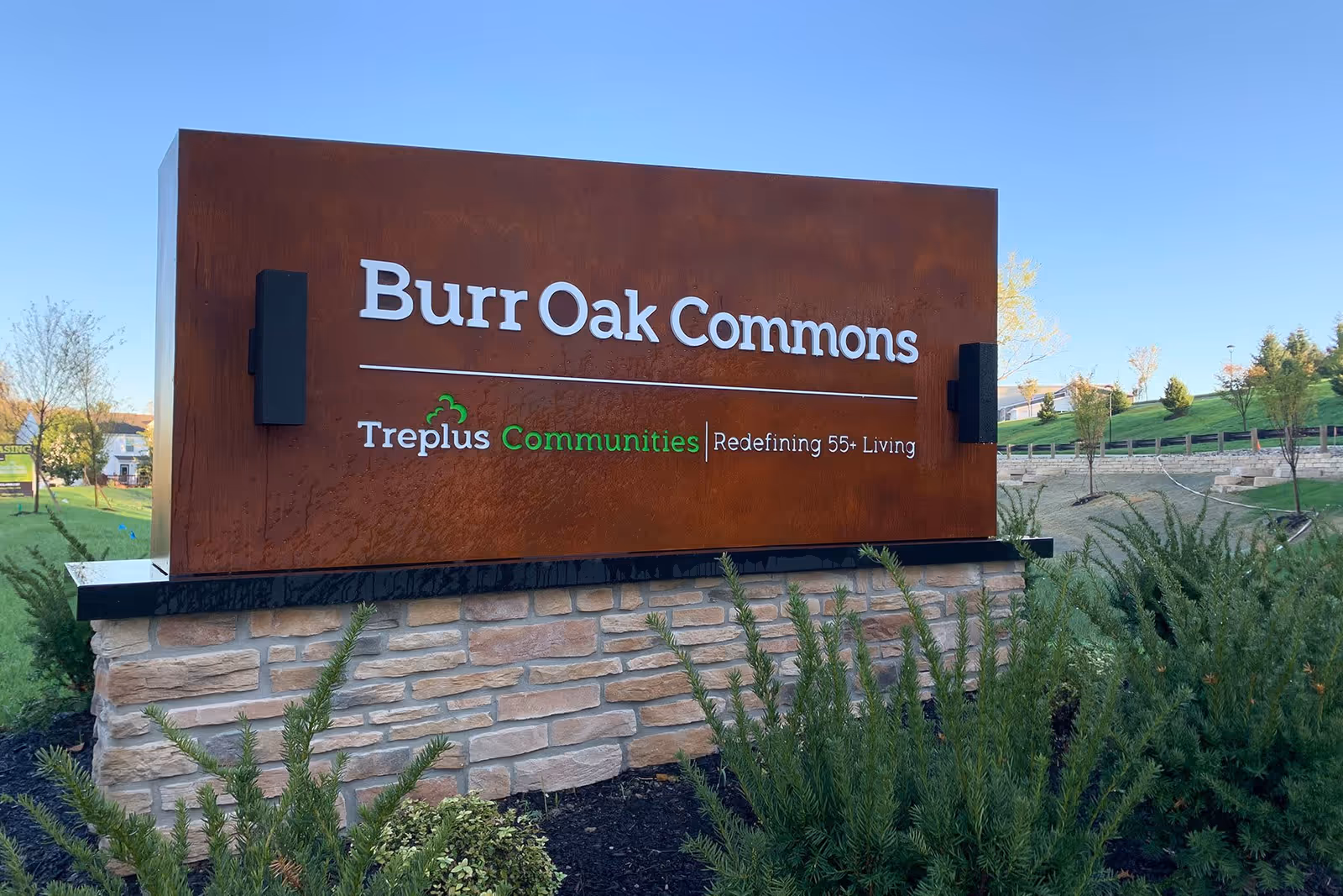 Outdoor sign for Burr Oak Commons, a community by Treplus Communities, with the tagline 'Redefining 55+ Living'. The sign is mounted on a stone base with greenery in front and a clear blue sky in the background.