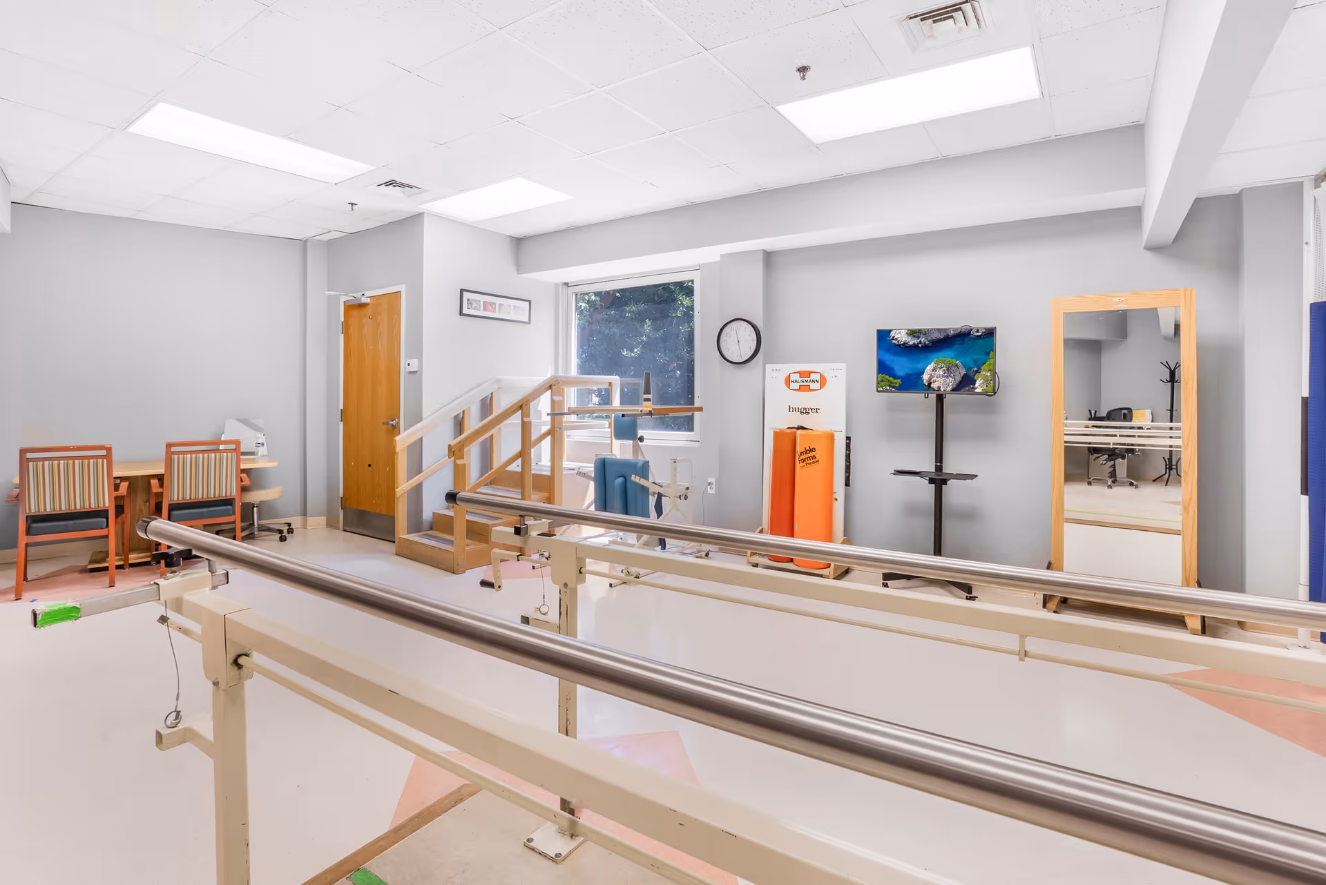 Physical therapy room with parallel bars for walking exercises, a small set of stairs with a handrail, a wall-mounted TV, a large mirror, and a desk with two chairs in the corner. The room has a window letting in natural light and light gray walls.