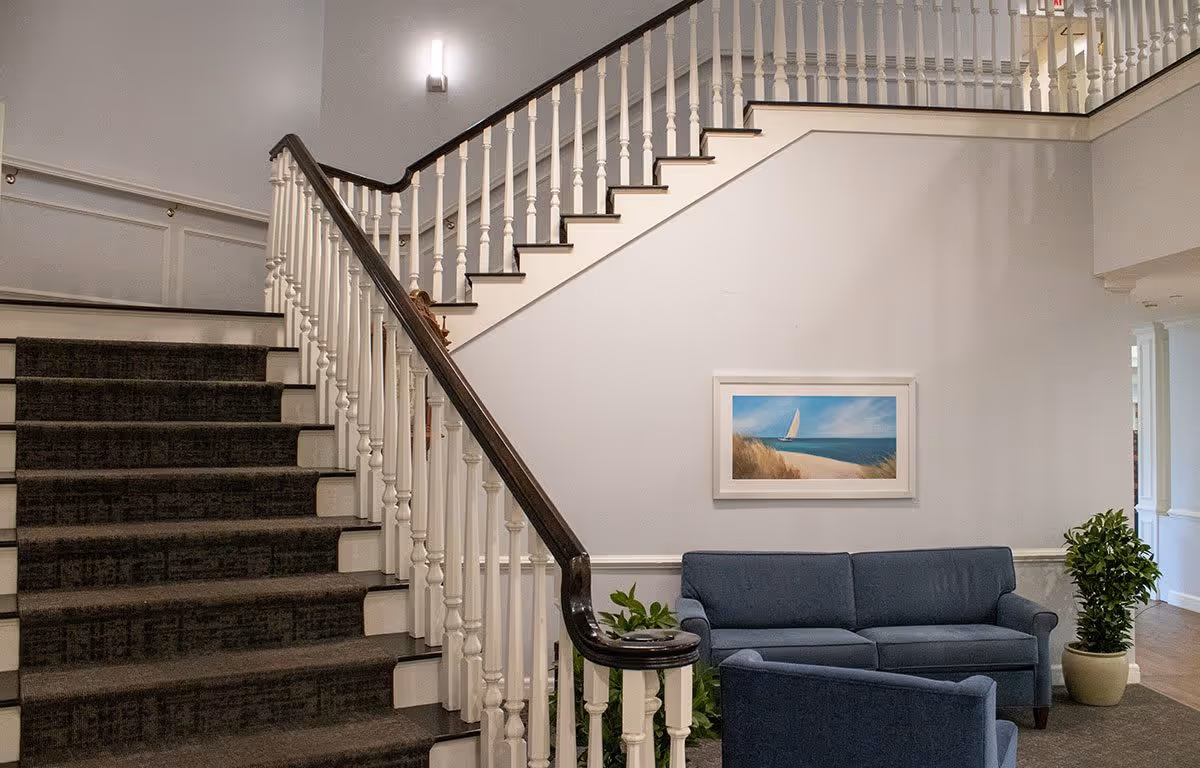 Interior view of a senior living facility showing a carpeted staircase with white railings and dark handrails. Below the staircase is a seating area with a blue sofa and a blue armchair, a potted plant, and a framed picture of a sailboat on the wall.