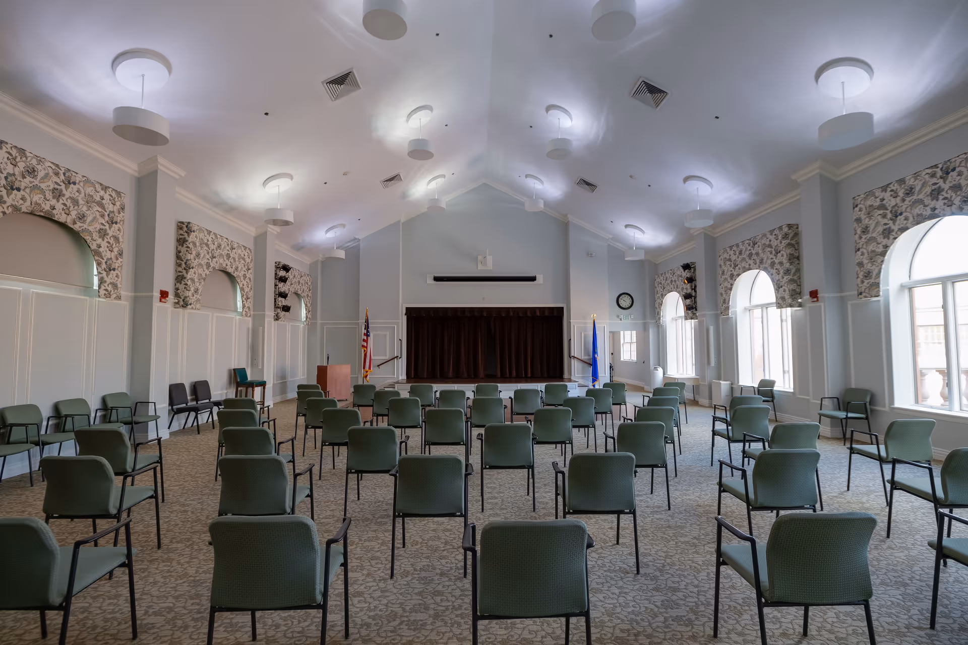 A large meeting or event room with rows of green chairs spaced apart facing a small stage with a closed curtain. The room has high ceilings with modern light fixtures, arched windows with floral curtains, and American and state flags on either side of the stage.