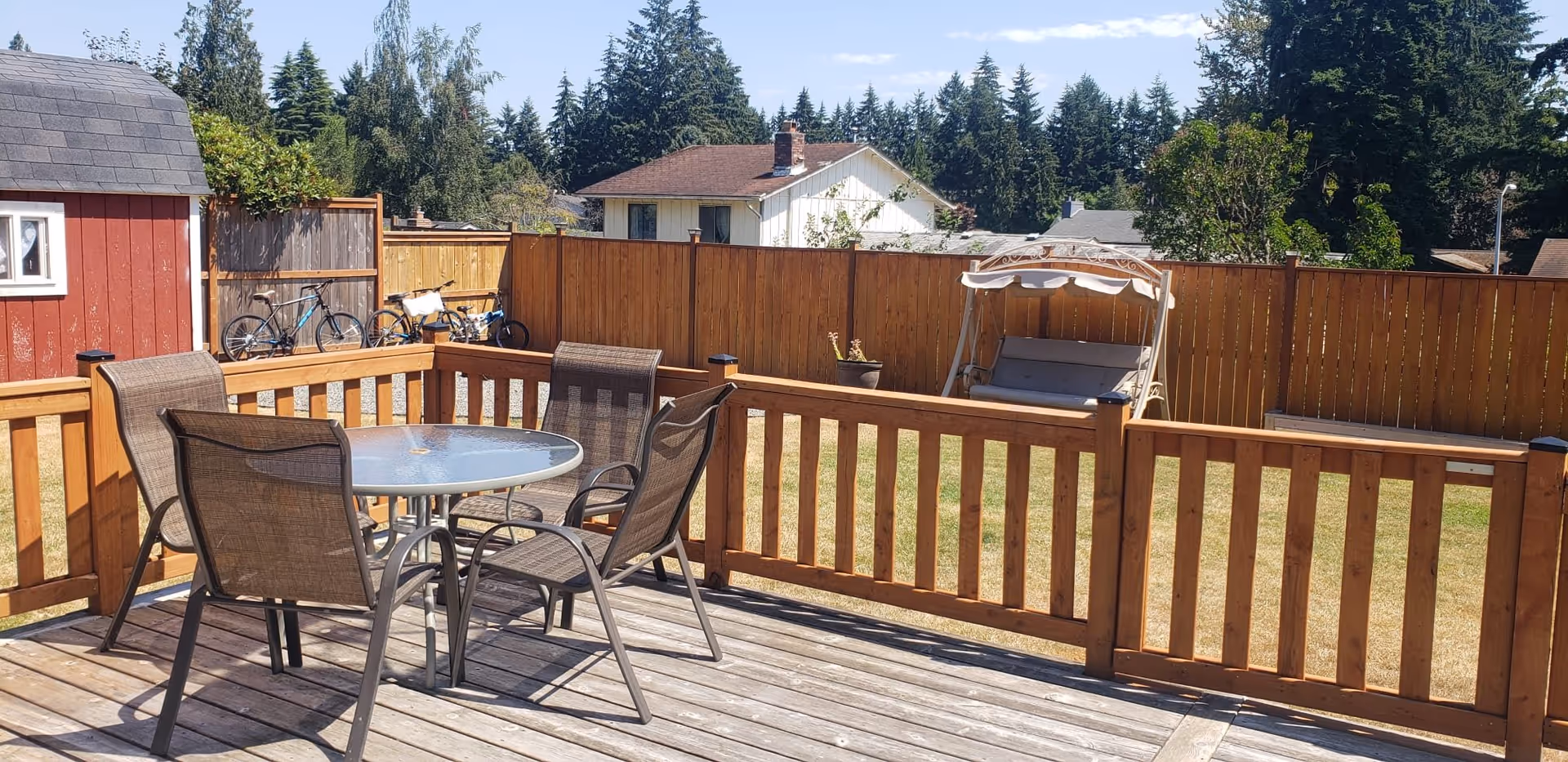 Outdoor wooden deck with a round glass table and four brown mesh chairs. Beyond the deck is a fenced yard with a beige cushioned swing seat, a red shed, and two bicycles leaning against the fence. Trees and houses are visible in the background under a clear blue sky.