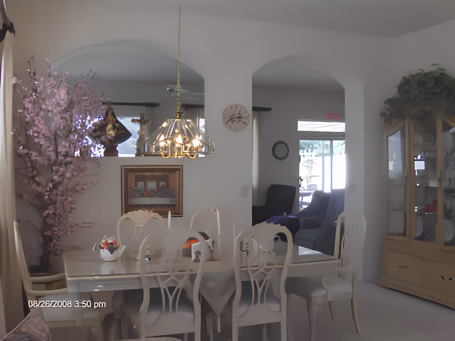 Interior view of a dining room with a white dining table and matching chairs. A chandelier hangs above the table. There is a large pink floral arrangement on the left side near a window with curtains. The room has white walls with two arched openings leading to another room with armchairs and a glass cabinet on the right side.