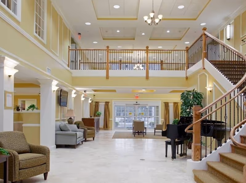 Spacious and well-lit senior living facility lobby with a grand staircase on the right, a black grand piano near the staircase, several armchairs and sofas arranged along the left wall, and large windows with curtains at the far end letting in natural light.