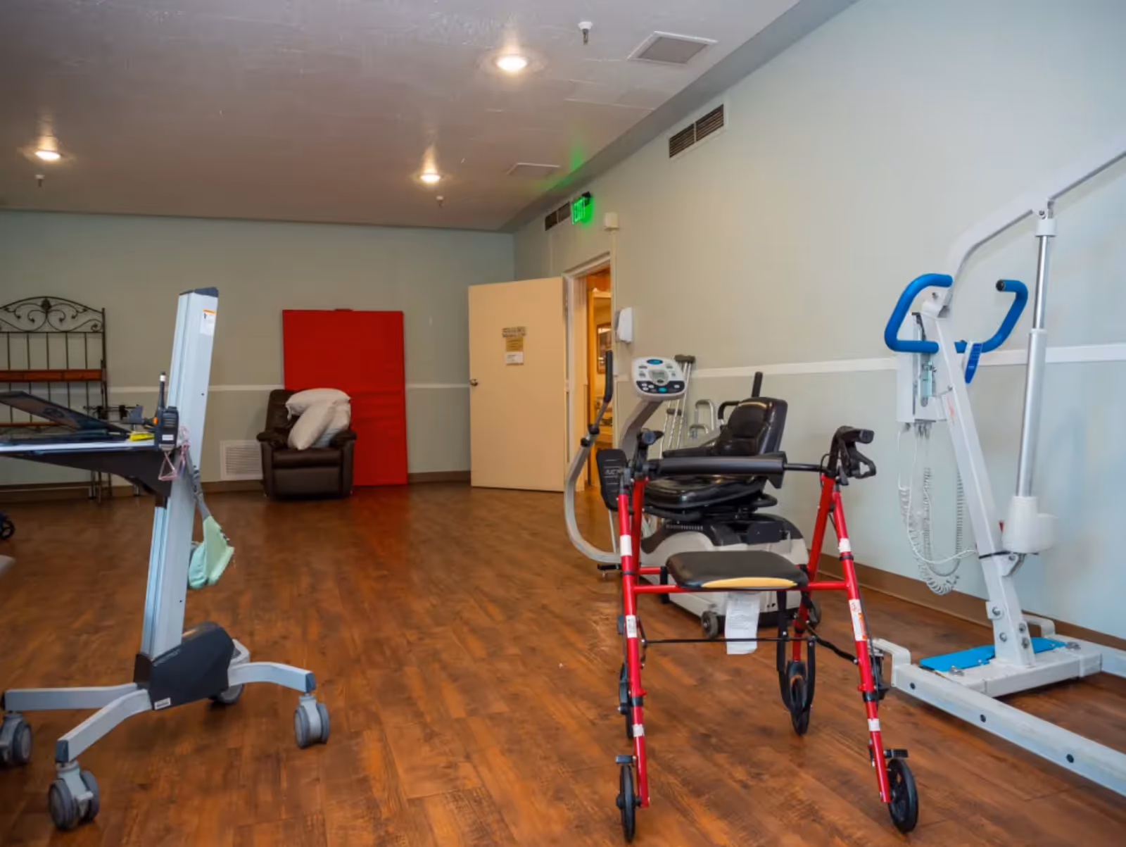 A rehabilitation room with wooden flooring featuring various physical therapy equipment including a red walker, a stationary exercise bike, and a patient lift. There is a brown recliner chair with pillows against the far wall, a red padded mat leaning against the wall, and an open door leading to another room.