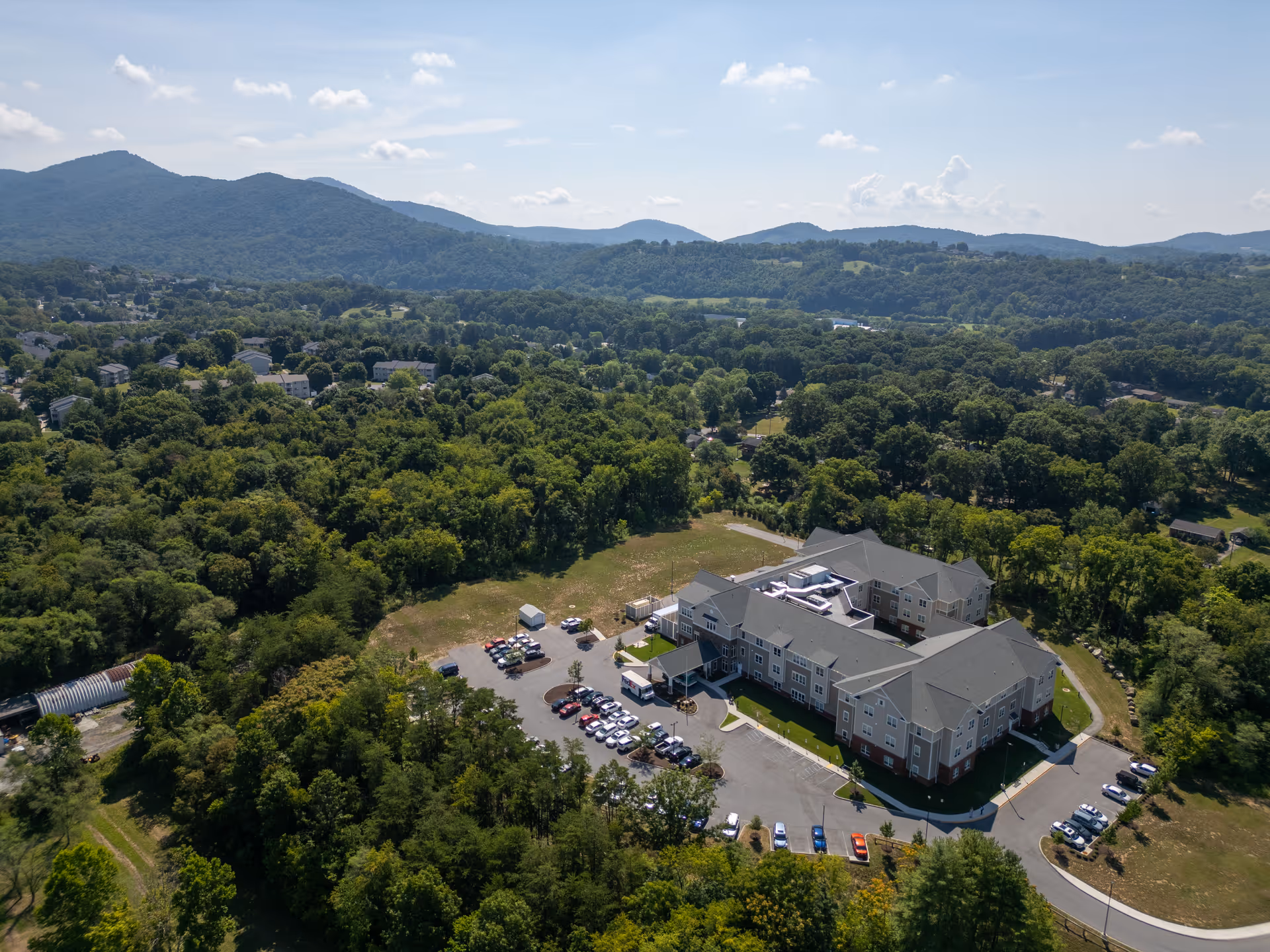 Aerial view of a multi-wing senior care facility with a parking lot surrounded by trees and hills in the background.