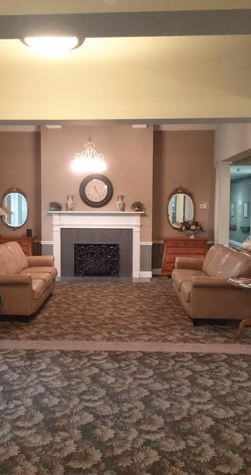 A cozy living room area with two tan leather sofas facing each other across a patterned carpet. In the center background is a white fireplace with decorative vases and plants on the mantel, a round clock above it, and a chandelier hanging from the ceiling. On either side of the fireplace are wooden dressers with oval mirrors and floral arrangements. The walls are painted in neutral tones.