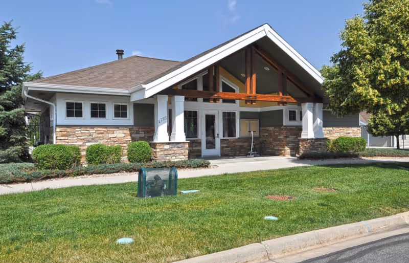 Exterior view of a single-story building with a peaked roof, stone and wood siding, and a covered entrance. The building is surrounded by green grass, shrubs, and trees under a clear blue sky.